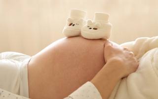 Schwangerer Bauch mit weißen, gestrickten Babyschuhen darauf, symbolisiert Schwangerschaft und Geburtshilfe in der Gynäkologie.