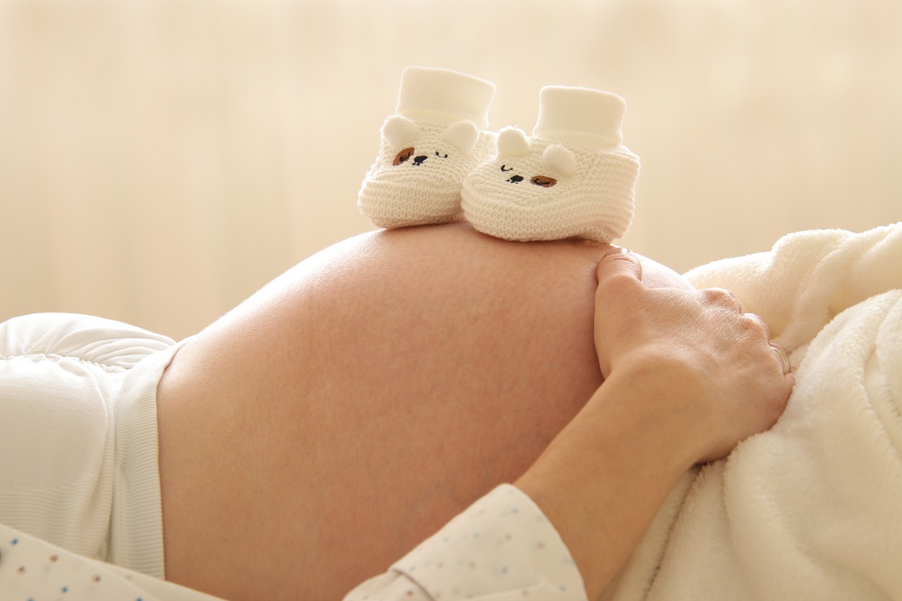 Schwangerer Bauch mit weißen, gestrickten Babyschuhen darauf, symbolisiert Schwangerschaft und Geburtshilfe in der Gynäkologie.